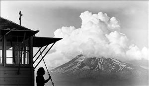 The silhouette of a person standing beside a small wooden structure looking out at a tall mountain with a thunder cloud covering its peak. 